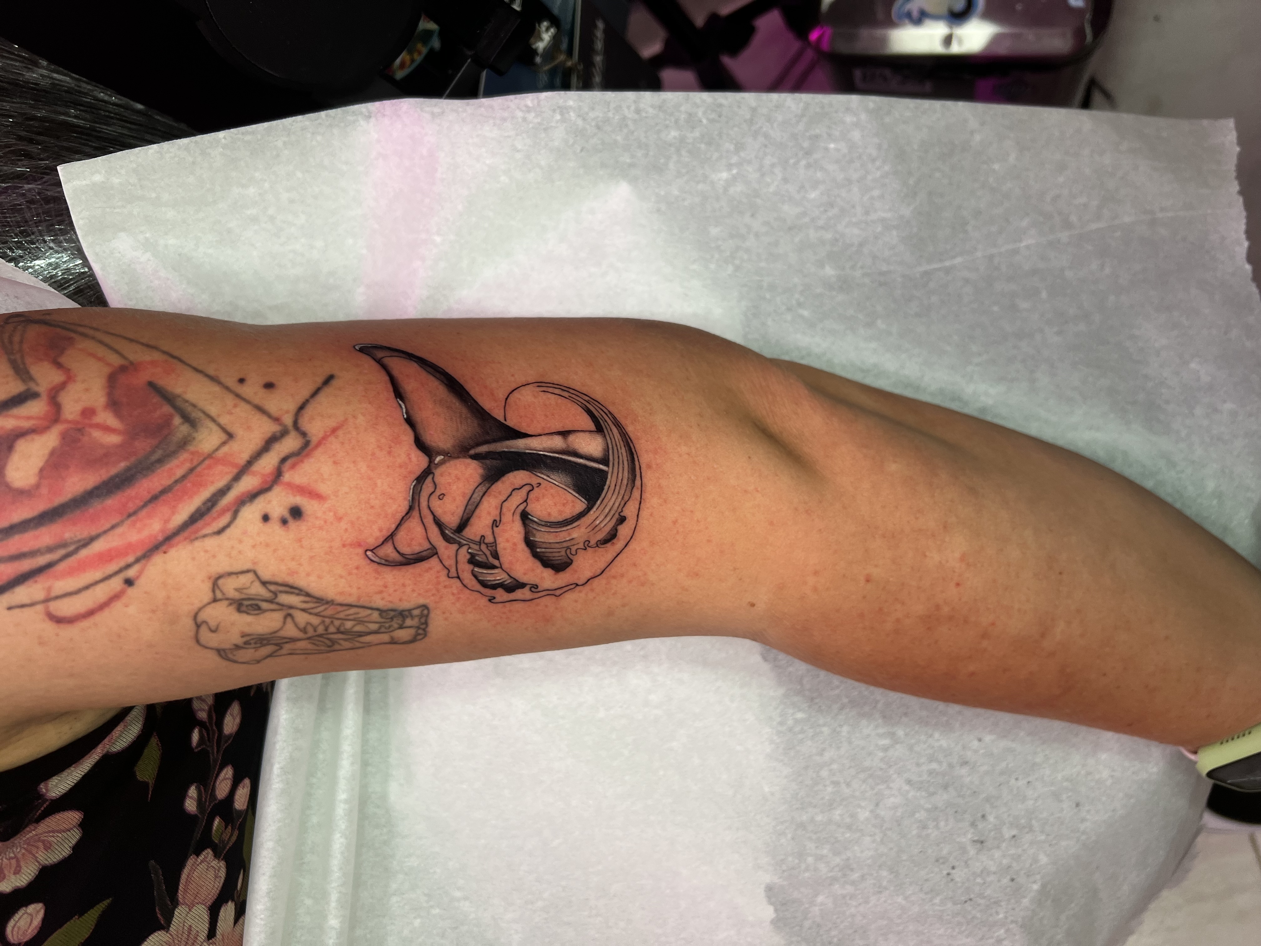 Es un tatuaje de estilo fine line con sombreado suave, donde la cola de una ballena emerge dentro de una ola que se enrolla formando un movimiento circular. La composición combina líneas limpias y del